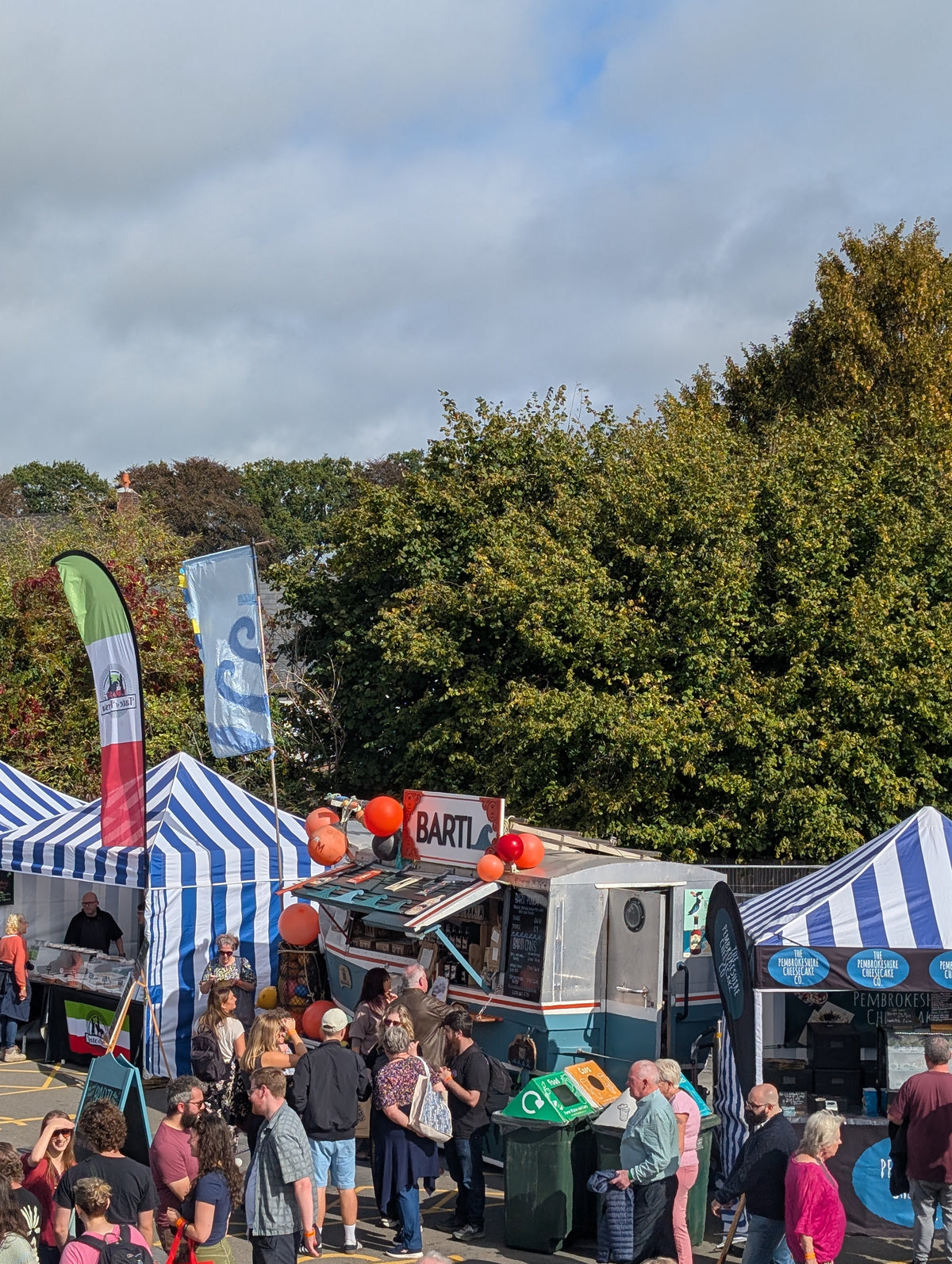 Outdoor market scene with blue-and-white striped tents, a food truck, people standing and walking, trees, and a cloudy sky.