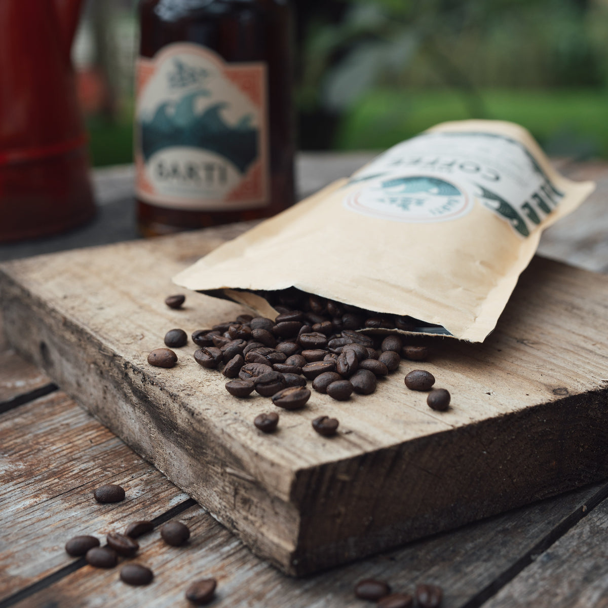 A kraft paper coffee bag lies open on a rustic wooden board with scattered roasted coffee beans, next to a dark bottle labeled BARTI and a red teapot in a softly blurred outdoor setting.