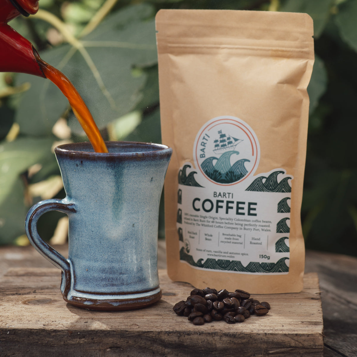A 150g bag of Barti Coffee stands on a rustic wooden board next to spilled coffee beans, with coffee being poured into a blue ceramic mug from a red pot in the background.