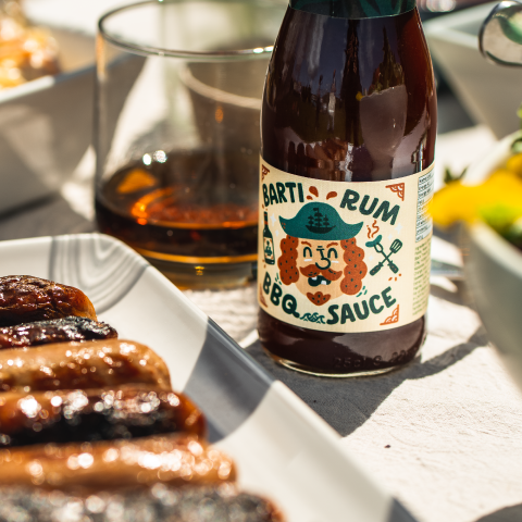 Best Spiced Rum BBQ Sauce, made with Barti Spiced Rum. BBQ Sauce bottle on a table with BBQ sausages and spiced rum in a glass